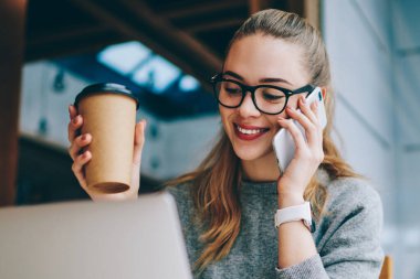 Kafeteryada kafein içeceği için eğlenen mutlu kız öğrenci ve akıllı telefon uygulaması aracılığıyla arkadaşınla akıllı telefon sohbeti, kafede dizüstü bilgisayarla oturan ve uygulamayla arayan neşeli hippi kız.