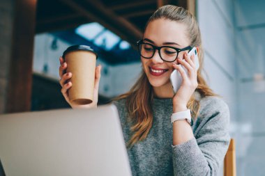 Kafeteryada kafein içeceği için eğlenen mutlu kız öğrenci ve akıllı telefon uygulaması aracılığıyla arkadaşınla akıllı telefon sohbeti, kafede dizüstü bilgisayarla oturan ve uygulamayla arayan neşeli hippi kız.