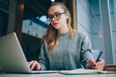 Kapalı alanlarda okuyan konsantre bir kız öğrenci netbook üzerinden okul ödevi için bilgi arıyor, gözlüklü hippi kız bilgisayarında ders kitabı okuyor.