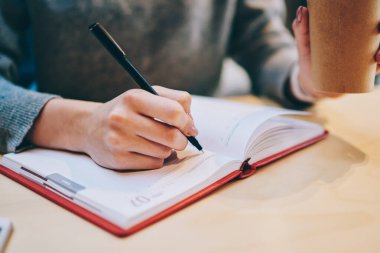 Kafeteryadaki kahve molası sırasında siyah kalem ve kırmızı not defteri kullanarak gelecek ay için planlama programının kesilmiş görüntüsü, ders kitabında bilgi notları alan kadın eline odaklanmış.