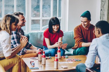 Kanepede oturup arkadaşlarıyla poker oynarken, akıllı telefondan gülümseyen ve kombinasyonu kontrol eden gündelik kıyafetli mutlu genç bir kadın.