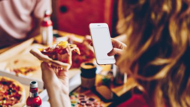 Yukarıdan gelen iştah açıcı pizza dilimi cep telefonuyla fotoğraf çeken bir grup çok ırklı insanla aynı masada yemek yiyor.