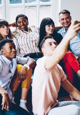 Yukardan gelen mutlu milenyum çeşitliliği öğrencileri günlük kıyafetleri içinde rahat modern apartman dairesinde buluşurken akıllı telefonlarıyla selfie çekiyorlar.