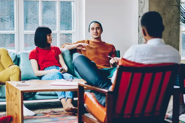 Modern bir dairede masanın yanında oturan ve etnik çeşitliliğe sahip bir grup arkadaşa ilginç bir hikaye anlatan gündelik kıyafetli genç bir adam.