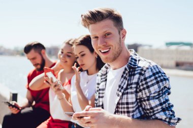 Yakışıklı, genç, gündelik kıyafetli, neşeli bir şekilde gülümseyen ve arkadaşlarıyla sette takılırken kameraya bakan ve modern akıllı telefonları karıştıran bir delikanlı. Teknoloji kullanan çağdaş insanlar.