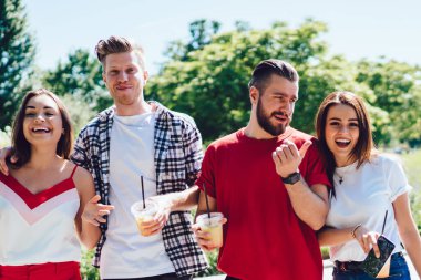 Güneşli bir yaz gününde parkta yürürken gülen ve taze soğuk içeceklerin tadını çıkaran modaya uygun kıyafetlerle bir grup genç.