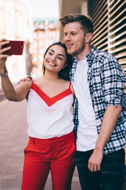 Yakışıklı, gülümseyen ve sarılan genç bir adam modern binanın yanındaki şehir sokağında dururken ve selfie için poz verirken. Mutlu milenyum çifti, telefon kamerasında kendilerini çekiyorlar.