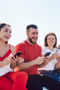 Gündüz kıyafetleri içinde neşeli bir şekilde gülümseyen ve akıllı telefonları karıştıran bir grup genç insan güneşli bir günde şehir sokaklarına karşı birlikte oturuyorlar. Arkadaşlar teknolojiyi açık havada kullanıyorlar.