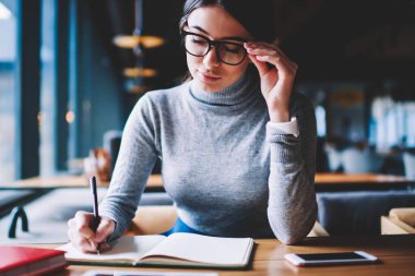 Konsantre ciddi kadın öğrenci ahşap masada oturma şık gözlükler ve defter yazma kompozisyon, gözlüklü istihbarat akıllı kadın, Not defteri kapalı bilgi analiz oturan oturuyor