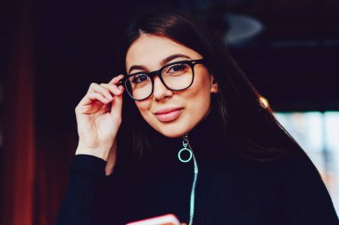 Kamera bakarak ve elinde akıllı telefon tutarak kahve dükkanında oturan pozitif gülümseyen hipster kız, gözlük poz ve yeni modern bilgisayar ile kafeteryada bekleyen arkadaş mutlu kadın