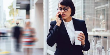 Resmi giyinmiş ciddi bir bayan yönetici elinde kahve tutarak akıllı telefondan konuşuyor. Kendine güvenen bir kadın patron, uygulamayla metropoldeki kentsel ortamlarda vakit geçiriyor.