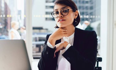 Gözlüklü, kendine güvenen kadın girişimcinin portresi şık kıyafetler giyip, finansal kafeteryada dizüstü bilgisayarla oturarak çevrimiçi çalışmak için vizyon düzeltmesi sağlıyor.