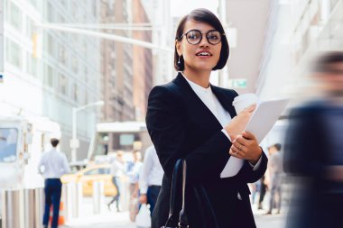 İş ortağıyla buluşmadan önce şehir ortamında duran ve iş molasından zevk alan görüş düzeltme gözlüklü pozitif kadın zarif takım elbiseli mutlu kadın ekonomist başka tarafa bakıyor.