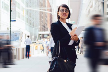 İş ortağıyla buluşmadan önce şehir ortamında duran ve iş molasından zevk alan görüş düzeltme gözlüklü pozitif kadın zarif takım elbiseli mutlu kadın ekonomist başka tarafa bakıyor.