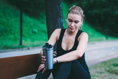 Aktif kadın Runner doğa üzerinde kardiyo eğitim mola zevk alüminyum Tumbler elleri ile ahşap tezgah üzerinde oturan ve germe egzersizleri hakkında düşünme, beyaz kadın açık havada dinlenme