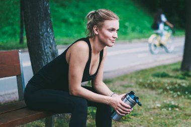 Kafkas kadın Runner el alüminyum termos ile ahşap tezgah üzerinde dinlenme alarak, siyah eşofman aktif sportif kız uzağa bakıyor ve doğa üzerinde iyi yapılmış sert eğitim iyi hissediyorum