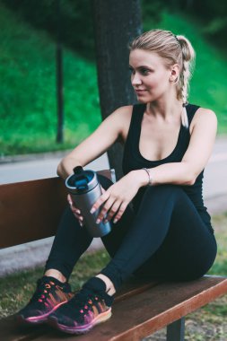 Kentsel ayar etrafında koşu sonra tezgah üzerinde dinlenme alarak güzel kadın, beyaz çekici kadın koşucu ahşap tezgah üzerinde oturan ve egzersiz için zaman içinde ellerde alüminyum Tumbler tutarak