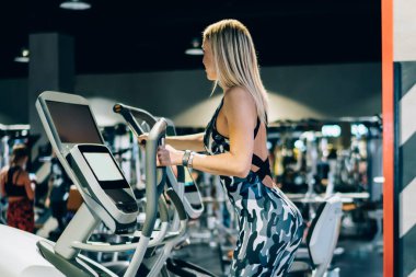 Sıska, atletik, sarışın, şık kamuflaj kıyafeti giyen ve spor salonunda egzersiz yapan bir kadının yan görüntüsü.