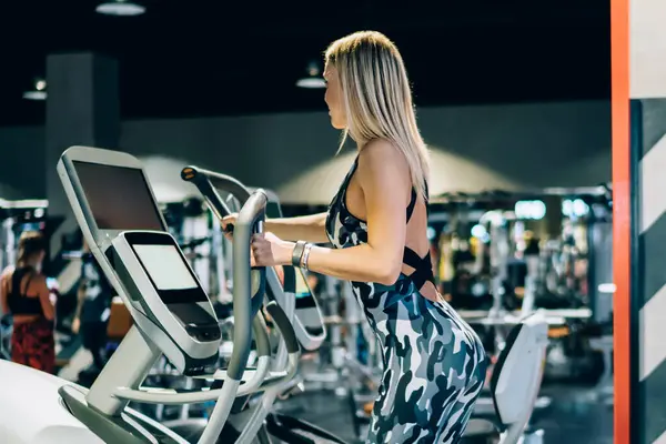 Sıska, atletik, sarışın, şık kamuflaj kıyafeti giyen ve spor salonunda egzersiz yapan bir kadının yan görüntüsü.