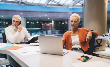 Modern bir ofiste çalışan kıdemli iş adamları, belgelerle telefonla konuşan bir adam, kadın ise dikkatle dizüstü bilgisayarına odaklanıyor. Takım çalışması, konsantrasyon, kurumsal çoklu görev kavramı