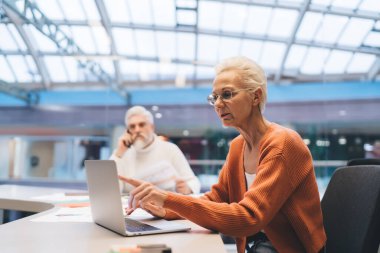 Modern ofisteki kıdemli iş adamları, arka plandaki adam telefondayken kadınların gülmesi ve dizüstü bilgisayarı göstermesi. Pozitif takım çalışması, iletişim ve iş yerinde keyif kavramı