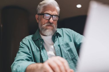 Gözlüklü, beyaz sakallı, laptop ekranına odaklanmış son sınıf öğrencisinin yakın çekimi. Beyaz bir tişörtün üzerine yeşil bir gömlek giyerek modern çalışma alanında konsantrasyon ve profesyonellik yayıyor.