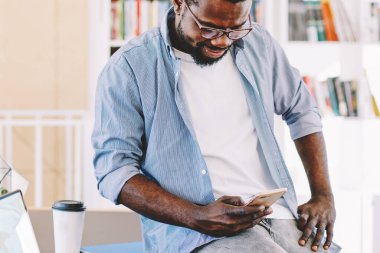 Aktif Afro-Amerikalı gözlüklü adam rahat bir şekilde masada dizüstü bilgisayar ve belgelerle çalışıyor ve cep telefonuyla ışık dolabında sörf yapıyor.