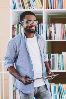 Gözlüklü zeki Afro-Amerikalı adam, kitaplıkların yanında duruyor ve aydınlık kütüphanede okumak ve aramak için tablet tutuyor.