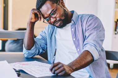 Sıradan giyinmiş ve gözlüklü Afro-Amerikan bir adam masada oturup geniş modern işyerinde taslak üzerinde düşünüyor.