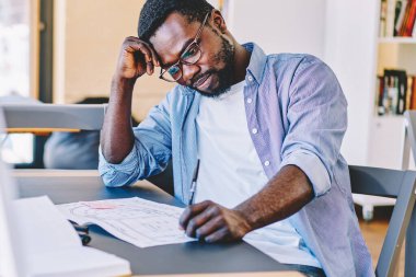 Sıradan giyinmiş ve gözlüklü Afro-Amerikan bir adam masada oturup geniş modern işyerinde taslak üzerinde düşünüyor.