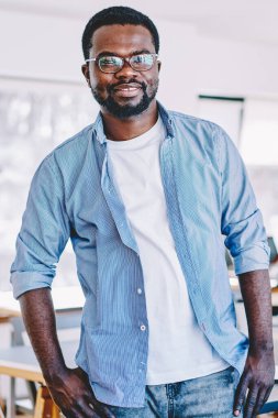 Rahat siyah adam gündelik giysiler içinde gülümseyen ve kameraya bakan geniş ofis molasında masaya yaslanan.