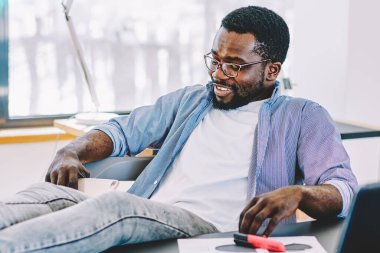 Rahat siyah adam gündelik kıyafetler ve gözlüklerle gülümsüyor ve modern işyerinde dinlenirken bacakları masanın üstünde tutuyor.