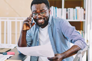 Gülümseyen, beyaz dişli, sıradan giyinmiş, cep telefonuyla arayan, elinde belge tutan, Afro-Amerikan bir erkek.