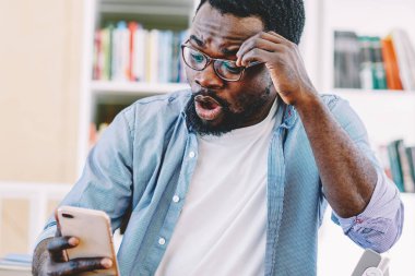 Şok olmuş, gözlüklü, sıradan giyinmiş, kafaya dokunmuş, cep telefonuyla bilgisayarı başında oturan Afro-Amerikan bir adam.