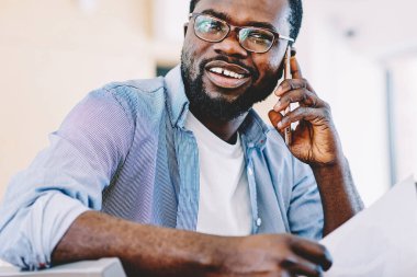 Beyaz dişli mutlu Afro-Amerikalı günlük giysiler içinde erkek cep telefonu kağıdını elinde tutarak beyaz koltukta oturuyor.