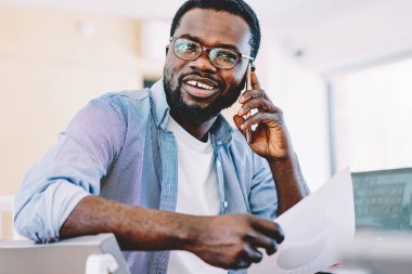 Beyaz dişli mutlu Afro-Amerikalı günlük giysiler içinde erkek cep telefonu kağıdını elinde tutarak beyaz koltukta oturuyor.