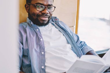 Siyah içerikli adam, duvarın yanındaki armut çuvalında oturmuş kameraya gülümsüyor ve elinde ilginç bir kitap tutuyor.