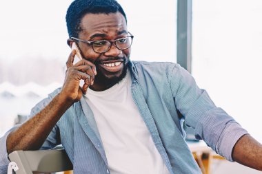 Tişörtlü ve gözlüklü siyah bir adam cep telefonuyla konuşuyor ve masada otururken ve dizüstü bilgisayar kullanırken gülümsüyor.