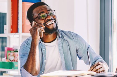 Düşünceli Afrikalı Amerikalı, gözlüklü, günlük kıyafetleri olan, telefonla konuşan, gülen ve evinde kağıt ve dizüstü bilgisayarla masada oturan biri.