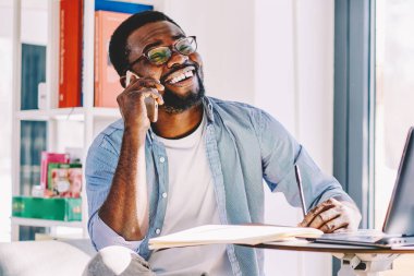 Düşünceli Afrikalı Amerikalı, gözlüklü, günlük kıyafetleri olan, telefonla konuşan, gülen ve evinde kağıt ve dizüstü bilgisayarla masada oturan biri.