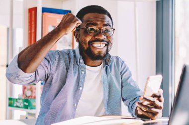 Gözlüklü, sıradan giyimli, neşeli Afro-Amerikan erkek. Zaferi kutlarken dizüstü bilgisayar ve kağıtlarla masada otururken evde akıllı telefon kullanıyor.