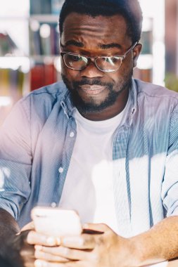 Gözlüklü ve günlük giysileri olan, güneşli bir günde evde otururken akıllı telefon kullanan, hayal kırıklığına uğramış yetişkin Afrikalı Amerikalı bir erkek.