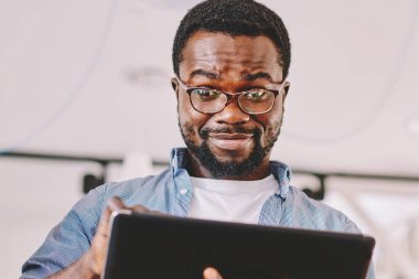 Aşağıdaki neşeli, Afrikalı, gözlüklü, serbest çalışan, elinde tabletle ayakta duran ve kameraya bakan adamdan.