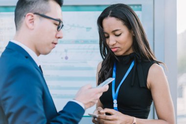 Zarif etnik kadın ve resmi üniformalı Asyalı adam demiryolu istasyonunda akıllı telefonlar kullanarak birlikte duruyorlar.