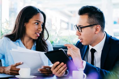 Genç Asyalı erkek ve Afrikalı Amerikalı kadın, kafede oturmuş, profesyonel bir toplantı yaparken, belgeleri tartışıyor ve tablet kullanıyor.