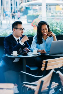 Genç, çok kültürlü bir kadın ve erkek kafeteryada oturmuş laptopla çalışıyor ve yeni iş projelerini tartışıyorlar.