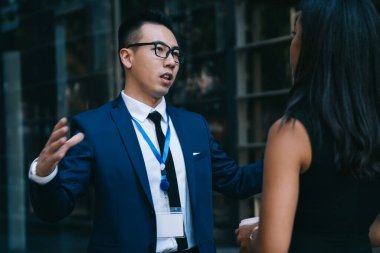 Takım elbiseli ve gözlüklü genç bir Asyalı iş konferansında etnik kadın iş arkadaşıyla konuşuyor ve kahve fincanlarıyla sokakta duruyor.