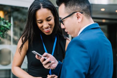 Genç, gülümseyen etnik kadın ve takım elbiseli Asyalı bir adam. İsim etiketi takıyor ve cep telefonu kullanıyor. Sokakta durup sohbet ediyorlar.