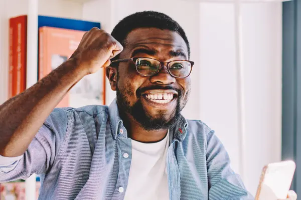 Gözlüklü, sıradan giyimli, neşeli Afro-Amerikan erkek. Zaferi kutlarken dizüstü bilgisayar ve kağıtlarla masada otururken evde akıllı telefon kullanıyor.