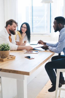 Aydınlık bir odada oturan mutlu çiftin yan görüntüsü ve sunulan etnik satış elemanlarının daire satın alma sözleşmesi önerirken gülümsemesi.